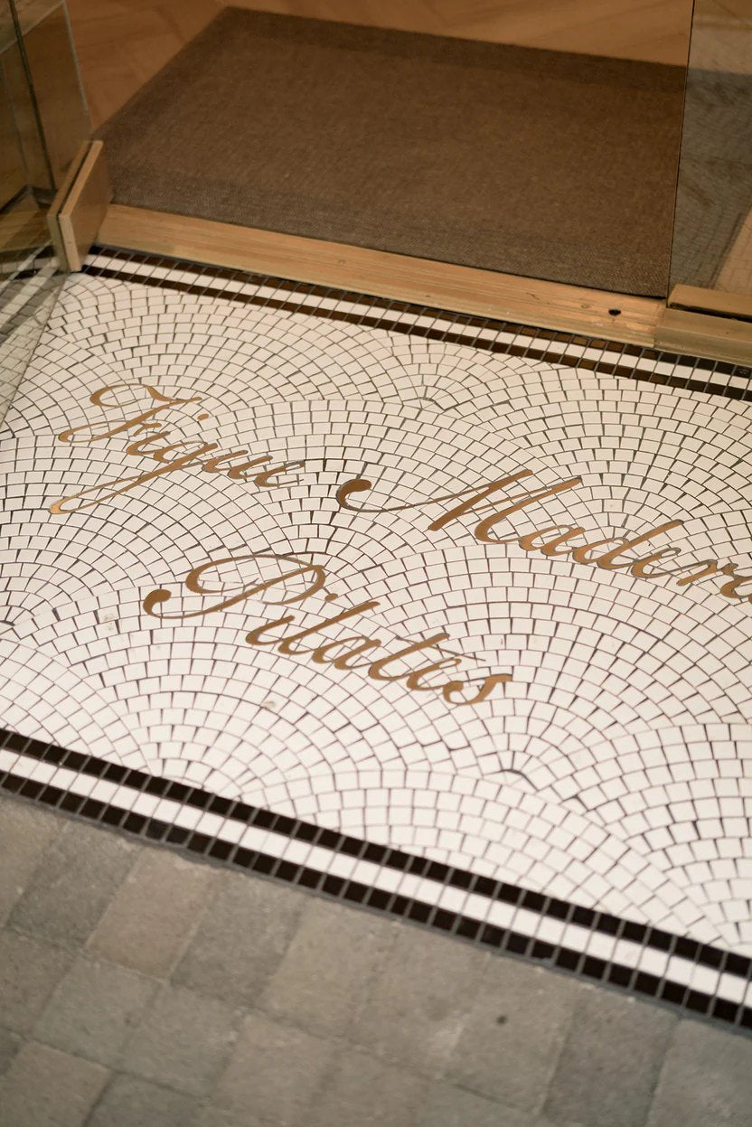 Gold metal script lettering on a white fan pattern mosaic at a pilates studio entrance
